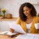 A woman sits at a kitchen table reviewing paperwork while holding a coffee mug, with a notebook and phone nearby in a bright, modern home.